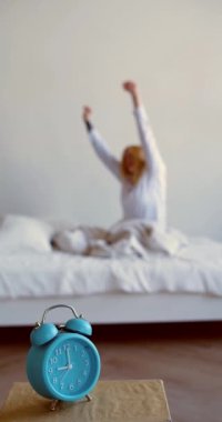 Ön planda bir çalar saatin dikey 4K görüntüsü ve bulanık sarışın bir kadın uyandı ve arka planda gerindi. Kollarını uzattı ve izin gününün tadını çıkardı..