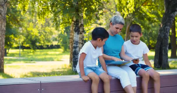 晴れた日に公園で2人の息子に本を読んでいるアジアの母親の心温まるシーン アウトドアで質の高い家族の時間を楽しむ — ストック動画
