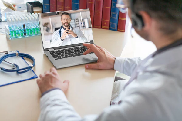 A doctor sits in their medical office and conducts a virtual consultation with a colleague using a laptop. The doctors are discussing a patients medical history and treatment options.
