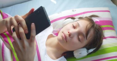 Genç kız kulaklıkla yatakta uzanıyor, akıllı telefonuyla arkadaşlarıyla çevrimiçi iletişim kuruyor. Gençlerin sosyal medyaya, çevrimiçi iletişim ve oyun bağımlılığı.