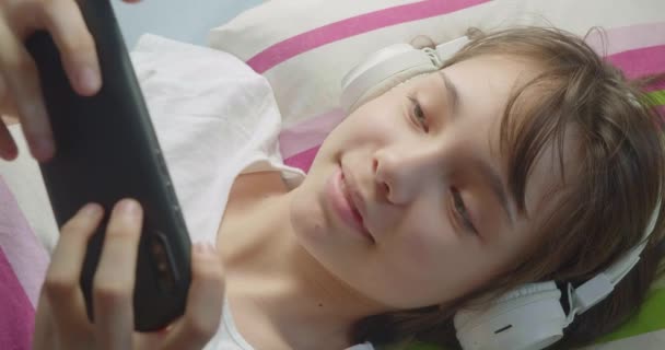 Nastolatka Całkowicie Zanurzona Grze Online Swoim Smartfonie Leży Łóżku Słuchawkami — Wideo stockowe