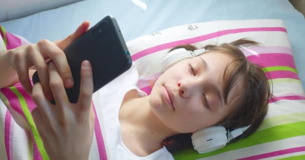 10代の女の子はヘッドフォンでベッドに横になり スマートフォンを通して友人とオンライン通信を行っています 若者のソーシャルメディアへの依存 オンラインコミュニケーション ゲーム — ストック動画