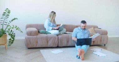 Kadın ve erkek evde çalışıyor. Evde dizüstü bilgisayarlarıyla kanepede oturuyor ve belgelerle çalışıyorlar. Belgeleri incele ve bilgisayardaki verilerle kontrol et. 60 fps 4k görüntü.