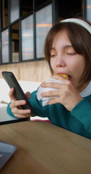 10代の女の子はファーストフードを食べながらスマートフォンの画面に吸収されました このような不健康な食品 特に若い人のために食べることの有害な影響 スローモーション垂直4K映像 — ストック動画