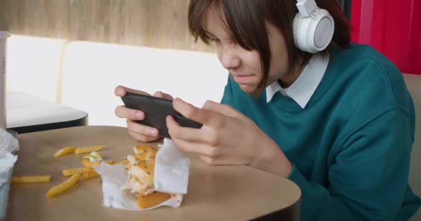 ファーストフードのカフェに座っている間 10代の女の子がスマートフォンの画面に吸収されます 彼女は彼女のスマートフォンでオンラインゲームをプレイしています デジタル世界に現実を脱出 スローモーション4K — ストック動画
