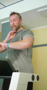 Antrenman sırasında kalp atış hızı kontrolü. Orta yaşlı bir adam koşu bandında koşar ve giderken nabzını kontrol eder. Dikey 4k görüntü.