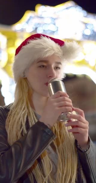 圣诞前夕 戴着圣诞礼帽的少女在户外喝着能量饮料 在节日灯火通明的情况下 年轻女人喜欢喝铝制的能量饮料 Verrtical Video — 图库视频影像