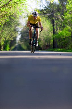 Güneşli bir günde yeşil ormanda asfalt yolda koruyucu miğfer ve bisiklet giyen bir sporcu. Düzenli egzersiz ve sağlıklı yaşam tarzı. Dikey fotoğraf.