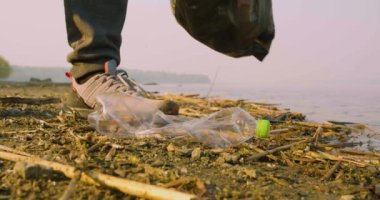 Çevreye önem vermek. Göl kıyısındaki çöpleri toplayın. Koruyucu eldivenli bir insanın yerden plastik bir şişeyi alırken yakın plan görüntüsü..