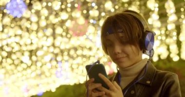 İnternette akıllı telefon sohbeti yapan genç bir kız. Noel arifesinde şenlik ışıkları altında sokaktaki genç kız. İnternette müzik dinliyor ve arkadaşlarıyla sohbet ediyor. 60 fps görüntü.