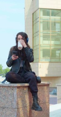 Şehir sokaklarında dinlenirken arkadaşlarıyla internette sohbet eden genç bir kadının dikey görüntüleri. Modern bir şehrin canlı ritmi ve çevrimiçi iletişimi uzaktan. Dikey görüntüler.