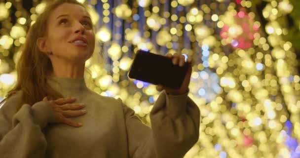 親戚や友人とのオンラインビデオ通話中の若い笑顔の女性のクローズアップビデオ 彼女はクリスマスライトの通りに彼女のスマートフォンを保持し 話しています スローモーション4K映像 — ストック動画
