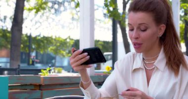 Kahve molası sırasında akrabalarla veya arkadaşlarla çevrimiçi görüntülü görüşme. Genç bir kadın kaldırımdaki kafede otururken akıllı telefonunu tutar ve konuşur. Yavaş çekim 4k görüntü.