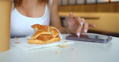 Genç bir kadın bir fast food kafede internetten iletişim kuruyor. Kırpılmış video 4K. Ivır zıvır yiyecekler ve sosyal ağlar gençliğin başlıca zararlı bağımlılarıdır..