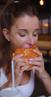 Kafede hamburger yiyen genç bir kadının yakın çekim görüntüleri. Fast food ve abur cubur modern toplumun bağımlılığıdır. Ağır çekim. Dikey 4k video.