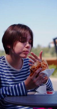 Kaldırımdaki kafede hamburger yiyen genç bir kızın yavaş çekim dikey 4K videosu. Parmaklarındaki lezzetli sosu yalıyor ve hamburgeri ısırıyor..
