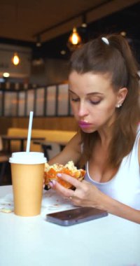 Bir kafede çizburger yiyen genç bir kadının dikey görüntüleri. Fast food ve abur cubur modern toplumun bağımlılığıdır. Yavaş çekim 4k görüntü.