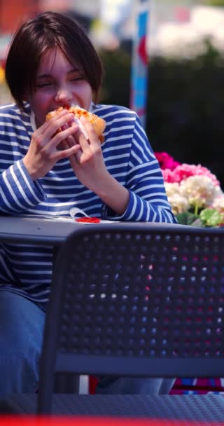 歩道カフェでチーズバーガーを食べている10代の女の子の垂直ビデオ スローモーション4K映像 — ストック動画