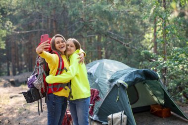 Anne ve kızı bir orman kampında selfie çekiyorlar. Çadırın yanında duruyorlar ve akıllı telefonlarıyla fotoğraf çekiyorlar. Genç neslin anneliği ve güveni..