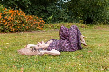 Güzel orta yaşlı bir kadın halka açık bir parkta çimlere uzanmış gülüyor.