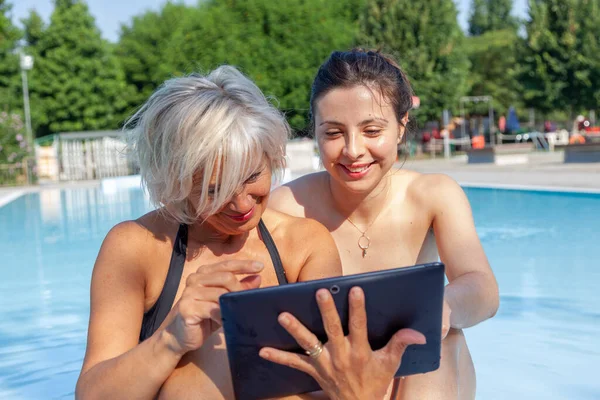 middle aged mom and young daughter dressing swimwear having fun with a tablet in a poolside - Concept of beautiful active people in summertime