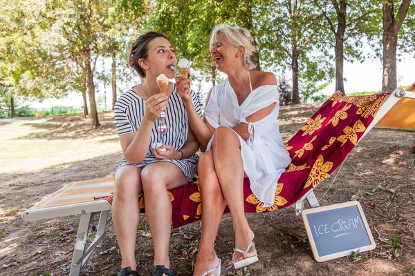 Modern mom and young daughter eating ice cream sitting on a deckchair in a water park on summer. Concept of beautiful people having fun in summertime