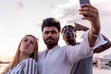 Küçük bir grup çok ırklı arkadaş akıllı telefonlu selfie çekerek eğleniyor. Yaz tatili konsepti nehirde giden bir teknede.