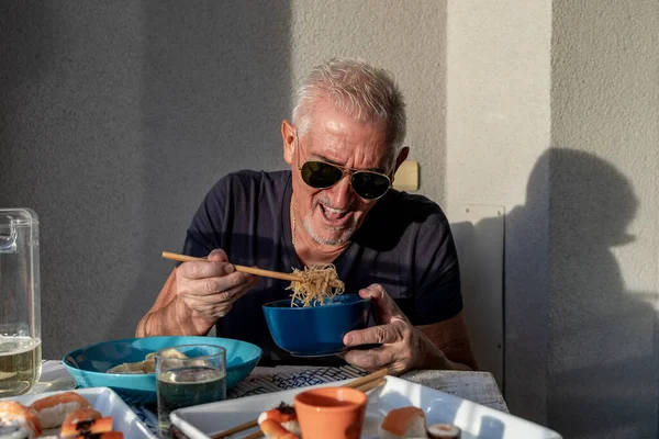attractive middle-aged man have fun while eating sitting at a table laid  Chinese take away food in front of gray wall outdoors