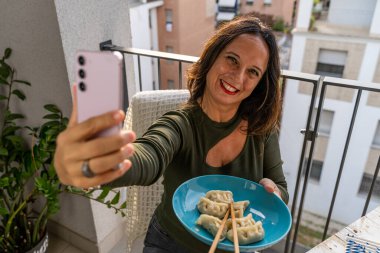 Güzel, orta yaşlı, romantik bir kadın selfie çekiyor ve gün batımında terasta yemek gösteriyor.