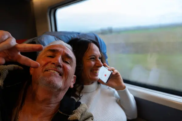 Happy middle aged couple taking a selfie sitting on a train having fun next to the window on vacation - Transportation and vacation concept.