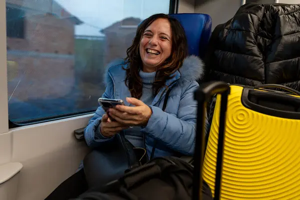pretty middle-aged woman traveling on the train sitting among the luggage next to the window using the smart phone - vacation and fun concept