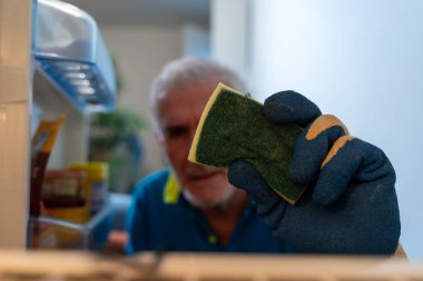 Orta yaşlı bekar adam buzdolabını neşeyle temizliyor. Ev aletinin içinden bakıyor.