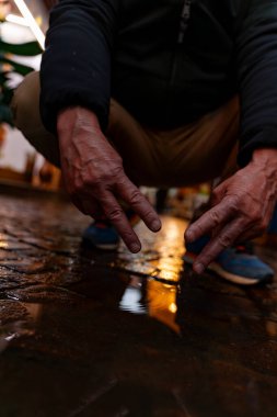 Mavi ayakkabılı bir adam bir su birikintisinin üzerine eğilip zafer işaretini gösteriyor.