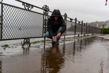 Orta yaşlı hippi yağmurun altında göl kenarında cep telefonlu bir su birikintisinin fotoğrafını çekiyor. Seyahat ve tatil kavramı.
