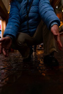 Mavi ayakkabılı kadın ayakları, akşamları bir şehir caddesindeki su birikintisine yansıyor.
