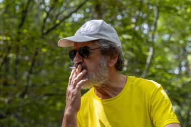 Olgun bir adamın dağ ormanlarında sigara içtiği portresi. Şapka, spor giysiler ve sırt çantası giymişti.