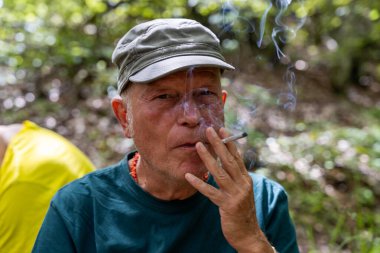 Olgun bir adamın dağ ormanlarında sigara içtiği portresi. Şapka, spor giysiler ve sırt çantası giymişti.