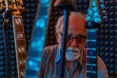 Orta yaşlı profesyonel gitarist gitar koleksiyonunun ortasında stüdyosunda poz veriyor.