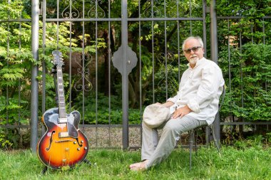 Orta yaşlı profesyonel gitarist evinin bahçesinde otururken etrafı gitaristlerle çevrili.