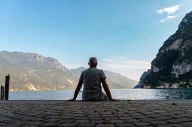 Orta yaşlı bir adam bir yaz günü Avrupa Gölü 'ne bakan manzaralı bir limanda oturuyor.