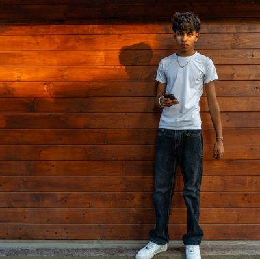 Yakışıklı genç gün batımında ahşap duvara yaslanarak cep telefonunu kullanarak eğleniyor.