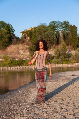 Gün batımında nehir kıyısında yürüyen kıvırcık saçlı güzel bir hippi kadının portresi.