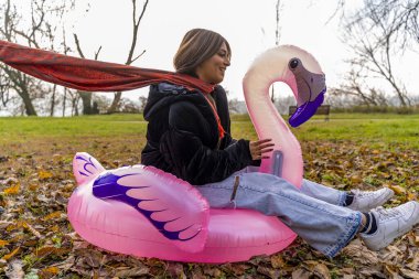 Genç ve güzel bir kadın kışın halka açık bir parkta şişme pembe bir flamingoya binerek eğleniyor.