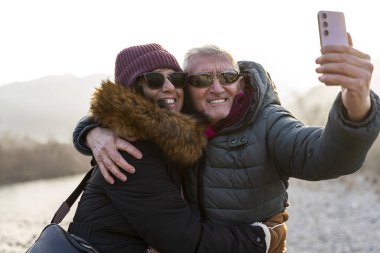 Güzel, mutlu, orta yaşlı, kış kıyafetleri içinde, rüzgarlı bir günde dışarıda selfie çeken bir çift.