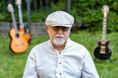 Orta yaşlı profesyonel gitarist evinin bahçesinde oturmuş gitar çalıyor. Etrafı gitarlarıyla çevrili.