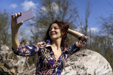 Çekici, güler yüzlü, kızıl saçlı bir kadın. Komik bir selfie çekiyor. Açık havada büyük bir kütüğün üzerinde oturuyor.