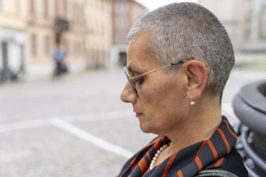 Açık havada oturan, tıraş olmuş gri saçlı orta yaşlı bir kadının profil portresi.