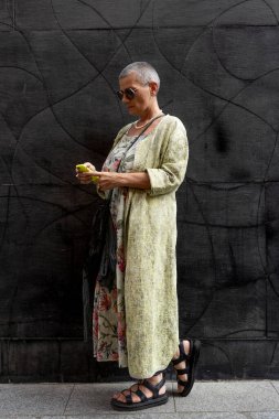 Tıraşlı, gri saçlı, siyah duvarın önünde cep telefonuyla mesajlaşan zarif, olgun bir kadın.