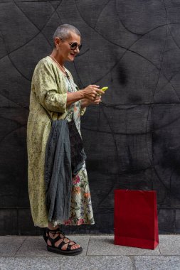 Tıraşlı, gri saçlı, siyah duvarın önünde cep telefonuyla mesajlaşan zarif, olgun bir kadın.