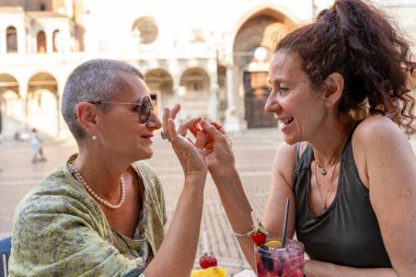 Güzel orta yaşlı lezbiyen çift, tarihi bir meydanda açık bir kafe masasında sevişiyor.